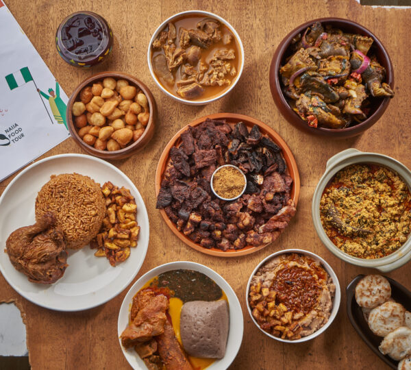 Assorted traditional Nigerian dishes including jollof rice with chicken, puff-puff, suya, egusi soup, pounded yam, and moi moi served on a wooden table with a Naija Food Tour flyer and a small Nigerian flag.