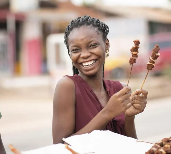 Woman holding freshly grilled Nigerian suya sticks, smiling at a Lagos street food stall.