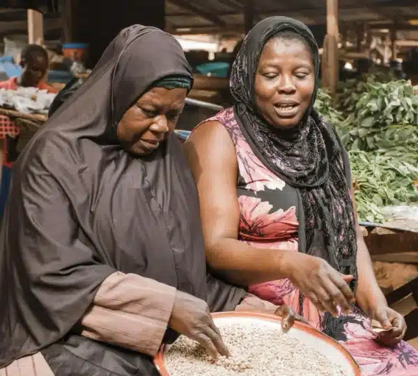 Two women smiling and shopping for vegetables at a busy market, reflecting the community spirit of local markets in Lagos.